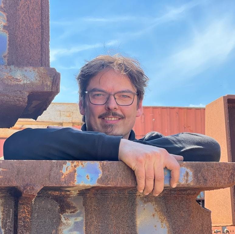 Foto van Gijs. Gijs is een witte man in de veertig met donkere ogen en kort bruin haar met slag. Gijs heeft een bruine snor en sikje. Op de foto draagt hij een blauw shirt met lange mouwen. Hij heeft zijn armen over elkaar geslagen en leunt op een ijzeren balk.