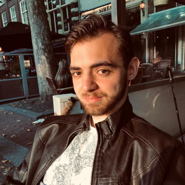 Foto van Nathan. Nathan is een jonge witte man met donkerbruine ogen, kort donkerbruin haar en een korte donkerbruine baard. Hij heeft een wit shirt en zwarte leren jas aan. Op de foto zit hij buiten op een terras.