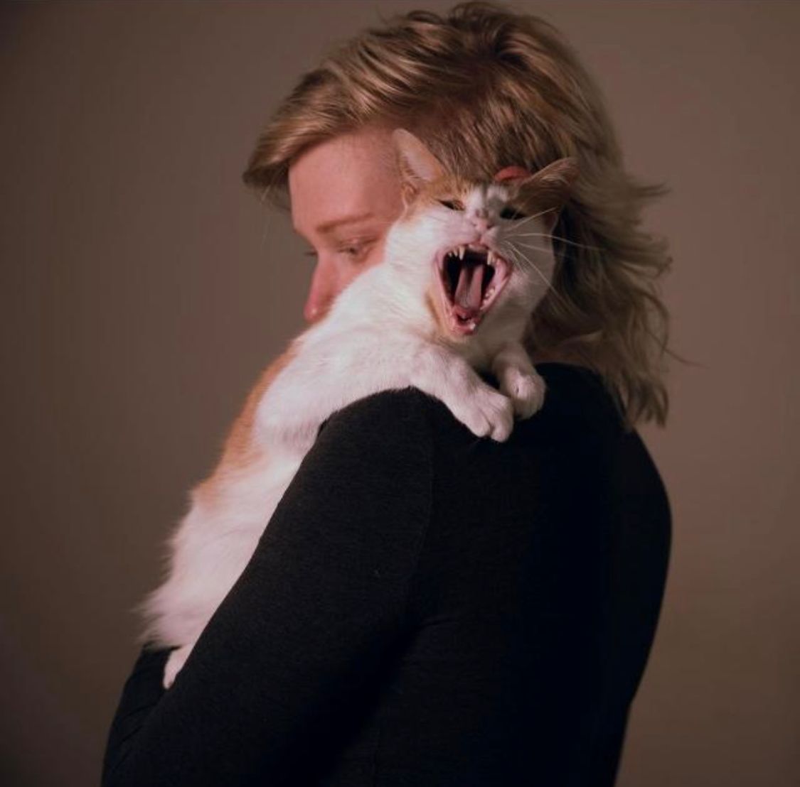 Foto van Tiva. Tiva is een jonge witte persoon met blauwe ogen en rossig blond haar in een mullet stijl. Op de foto is Tiva in het zwart gekleed, staat hen voor een bruine achtergrond en draagt hen zwarte kleding. Op haar schouder ligt een oranje witte kat, die naar de camera gaapt.